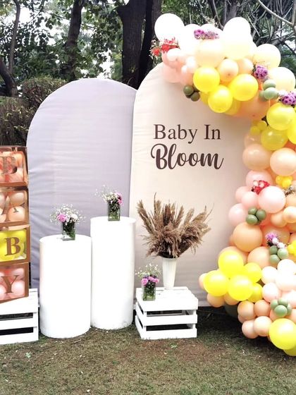 "Baby in Bloom" is such a beautiful theme for a baby shower. This outdoor setup features a lovely balloon arch in pastel yellows and pinks, elegant white pedestals, and custom "BABY" blocks, all set against a lush garden backdrop.