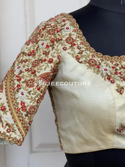 A beautiful off-white bridal blouse with elbow-length sleeves. The sleeves are heavily embroidered with a wedding procession theme, including elephant and floral motifs in red, white, and gold thread.
