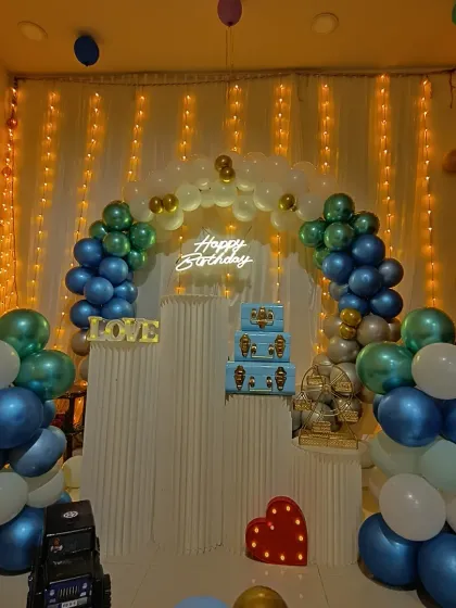 A birthday setup featuring a blue, green, and white balloon arch against a wall of fairy lights. The decor includes plinth stands, a light-up heart, and "LOVE" letters.