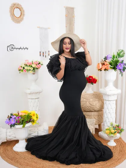 A stylish studio portrait of a mother-to-be in a black mermaid-style gown and a wide-brimmed hat, surrounded by flowers.