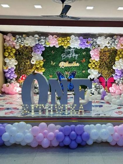 A butterfly-themed stage with a green turf backdrop and pastel balloon arch. The large 'ONE' letters and butterfly details create a beautiful garden-like setting.