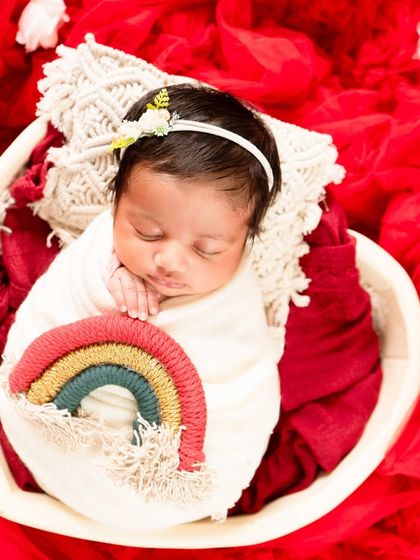 A newborn rests in a heart-shaped basket, holding a small rainbow prop against a vibrant red background. This setup is full of color and love.