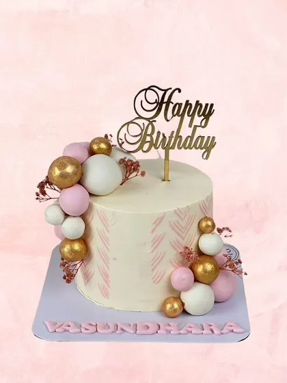 A chic and modern birthday cake for Vasundhara. The clean white frosting is accented with a subtle pink pattern and a cascade of pink, white, and gold spheres.