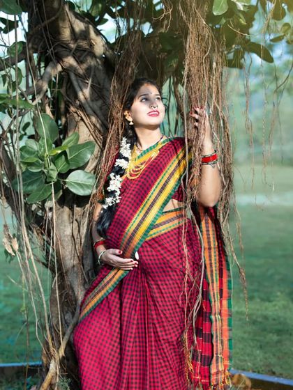 A breathtaking portrait of a mom-to-be in a traditional checkered saree. The golden hour light filtering through the banyan tree roots creates a magical, almost divine, atmosphere.