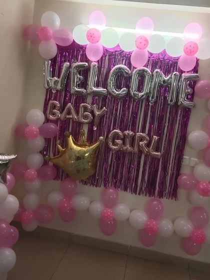 A 'Welcome Baby Girl' decoration with a pink and white theme. The setup features a balloon backdrop against a tinsel curtain and a crown foil balloon.