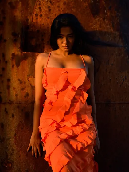 A full-length shot showcasing the vibrant orange dress against the rustic, textured background. The lighting is dramatic and warm.