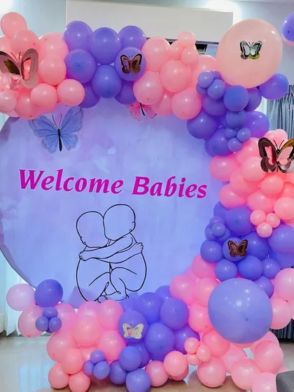 Celebrating the arrival of twins with this special "Welcome Babies" decoration. The setup includes a custom backdrop with a beautiful illustration, surrounded by a pink and purple butterfly themed balloon arch.