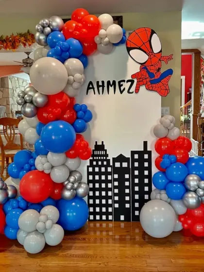 A Spider-Man themed birthday party setup. This decoration includes a city skyline backdrop, a Spider-Man cutout, and a balloon arch in the classic red, blue, and silver colors.