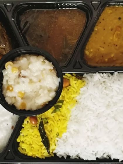A festival meal box with a variety of items. It includes lemon rice, white rice, curd rice, dal, sambar, sweet pongal, and some fried snacks.