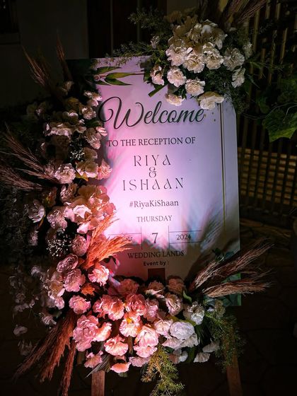 A beautifully styled welcome sign for a reception. The sign is adorned with an arrangement of white and blush flowers and pampas grass, setting an elegant and rustic-chic tone.