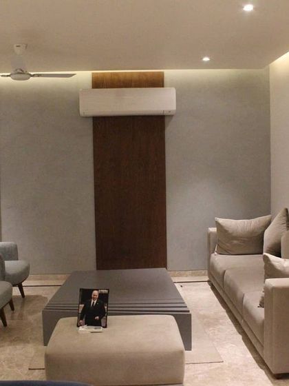 This view of the Hyderabad living room showcases the clean lines and balanced layout. A textured gray wall is complemented by a dark wood panel, creating a subtle feature behind the seating area. Cove lighting provides a soft, ambient glow throughout the space.