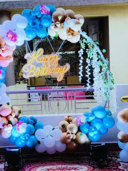 A beautiful floral and balloon decoration for a first birthday. This setup combines a circular balloon arch with hanging floral elements and a 'Happy Birthday' neon sign.