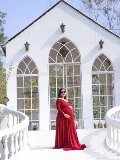 A beautiful solo portrait of a mother-to-be in a red gown, standing on a white bridge in front of a charming chapel-like building.