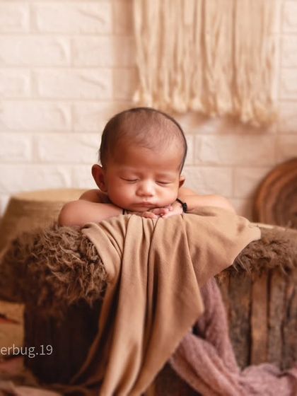 This newborn is posed sleeping on top of a textured log prop. The background features a white brick wall, macrame art, and dried pampas grass for a complete boho look.