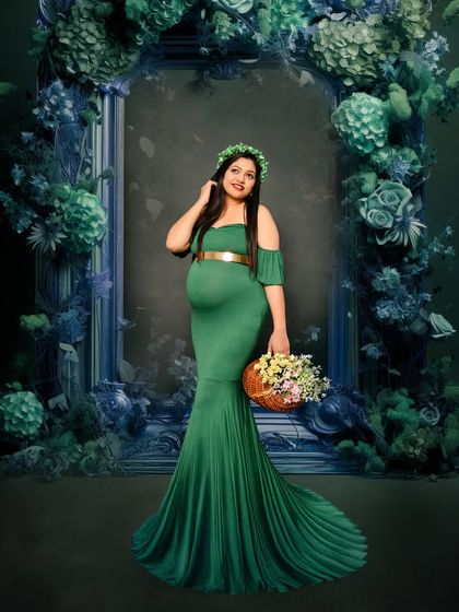 A beautiful solo portrait in a classic green gown. The mom-to-be is framed by a vintage-style mirror adorned with deep blue and green florals, creating a rich, moody atmosphere.