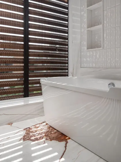 Sunlight streams through wooden blinds, casting beautiful shadows across the marble floor and freestanding bathtub in this luxurious master bathroom.