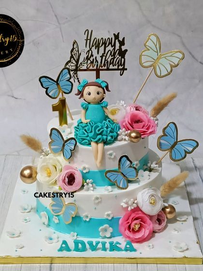 A lovely two-tier cake for a first birthday, decorated with fresh blossoms and butterflies. This pineapple cake features a girl figurine and a beautiful teal and white color scheme.
