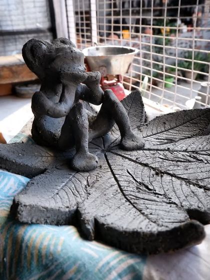 A whimsical sculpture of a figure sitting on a large leaf, created during a Raku workshop. The piece is shown here in its raw clay state before firing.