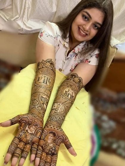 A happy bride showcasing her detailed portrait mehendi. The design includes figures of the bride and groom, along with architectural elements like jharokhas.