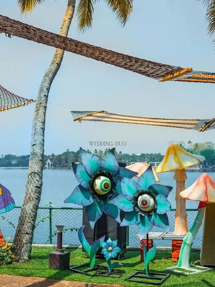 A whimsical, festival-inspired Mehendi by the lake. The decor features quirky, oversized installations like googly-eyed flowers and colorful mushrooms, with hammocks hanging from palm trees.