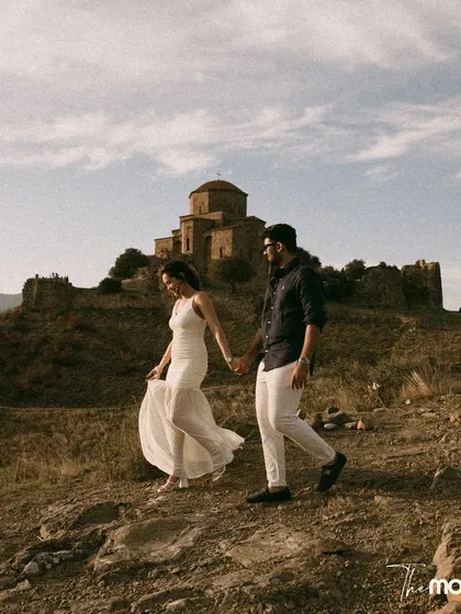 A couple walks hand-in-hand through the rustic, hilly landscape of Georgia, with a historic monastery in the background. The warm, earthy tones of the photo give it a timeless, romantic feel.