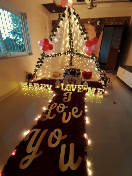 A full view of a romantic proposal or anniversary setup. A path of rose petals and lights leads to a cozy canopy tent, with 'I Love You' spelled out on the floor.