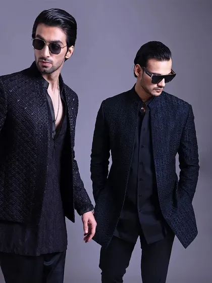 Two models showcasing all-black Indo-Western ensembles. The textured jackets and sharp tailoring create a look of modern power and poise.