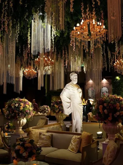 A luxurious lounge seating area at a reception, featuring classical statues, plush sofas, and dramatic overhead floral and light installations.