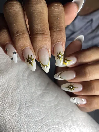 A celestial-themed design with a milky white base, gold starbursts, and rhinestone accents on stiletto nails.