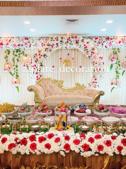 A beautiful baby shower setup with a floral arch and hanging decorative elements. The stage is filled with a complete arrangement of traditional plates, dolls, and a beautiful sofa.