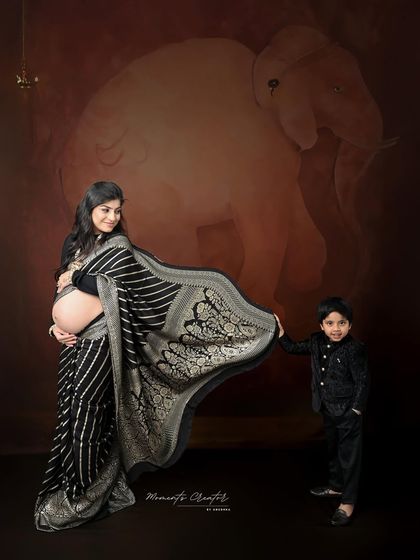 A sweet moment between mother and son. The older sibling proudly holds his mother's saree, set against a traditional backdrop with an elephant motif, symbolizing strength and family.