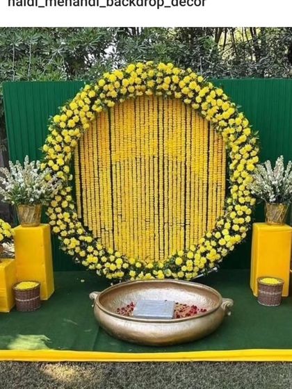 A classic Haldi backdrop for outdoor functions. This setup features a large circular frame filled with strings of yellow marigolds, a green panel background, and a traditional brass urli in the center for the ceremony.