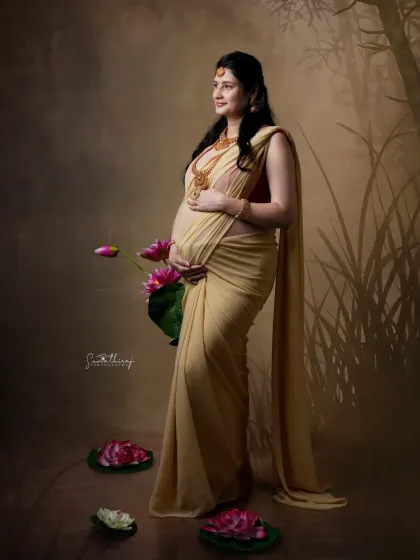 A full-length portrait of a mother-to-be in a flowing yellow saree, standing amidst lotus flowers. The composition is elegant and artistic, with a focus on her graceful posture.