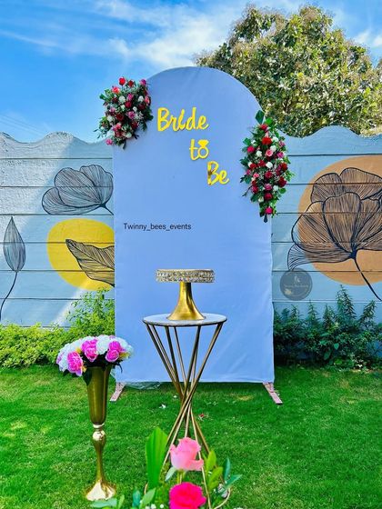 A simple and clean backdrop for a "Bride to be" party. The light blue arch is accented with small floral bouquets, providing a perfect canvas for a cake display in a garden setting.