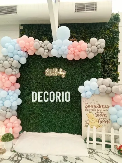 This baby shower setup uses a lush green hedge wall as a backdrop, creating a garden party feel. The "Oh Baby" sign and the pastel pink, blue, and grey balloon arch add a soft, celebratory touch.