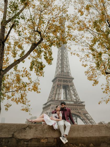 A romantic portrait of a couple sitting on a wall in Paris, with the Eiffel Tower as their backdrop. The autumn colors of the trees add warmth to this classic pre-wedding scene.