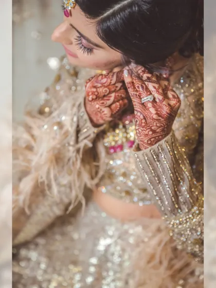 A detailed shot of the bride putting on her earrings. This close-up captures the intricate mehendi, the sparkling diamond ring, and the stunning details of her sequined and feathered lehenga.