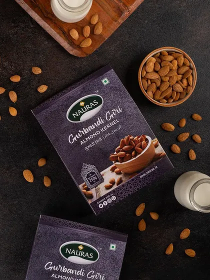 A top-down shot of Nauras Gurbandi Giri almond packaging on a dark, textured surface. The styling with bowls of almonds and milk suggests different ways to enjoy the product.