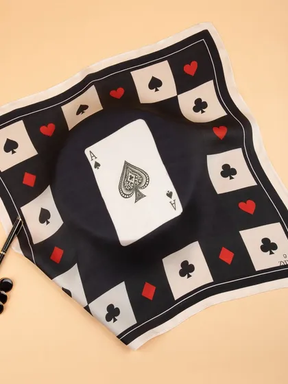 A creatively styled shot of the ace of spades pocket square, draped over a hat with a pen and game pieces to create a narrative.
