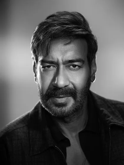 A black and white portrait of Ajay Devgn, styled for the film 'Shaitaan'. The look is understated yet intense, featuring a simple jacket and polo shirt to create a serious and brooding character persona.