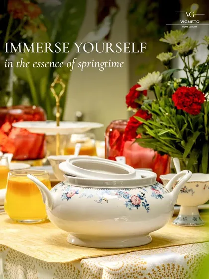 A lifestyle campaign image for Vigneto, featuring their floral serving dish on a vibrant, food-laden table. The text overlay "Immerse yourself in the essence of springtime" perfectly complements the visual story.