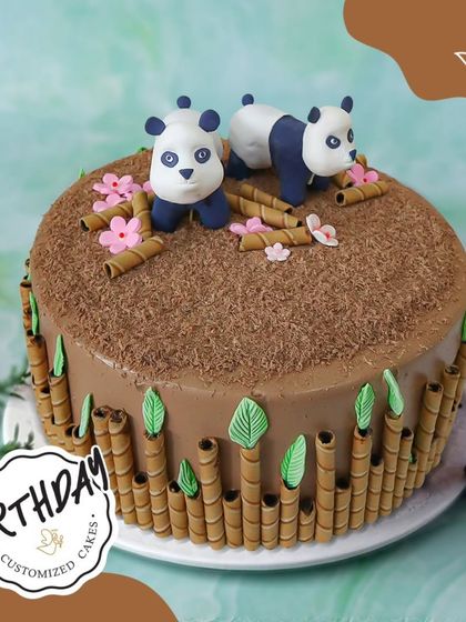 A delightful chocolate panda cake. This design features two cute pandas playing on a bed of chocolate shavings, surrounded by wafer stick bamboo.
