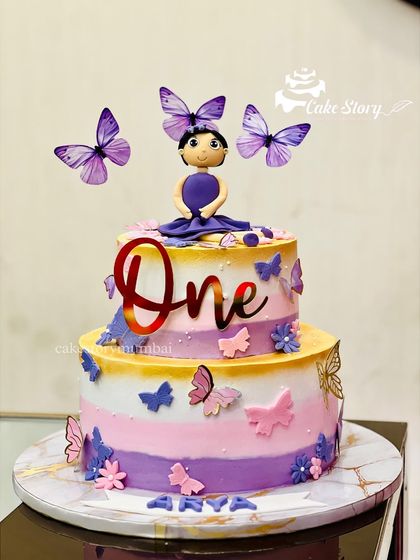 A vibrant butterfly-themed cake for a first birthday. This two-tier cake uses a colorful palette of purple, pink, and yellow, decorated with a fondant girl topper and lots of pretty butterflies.
