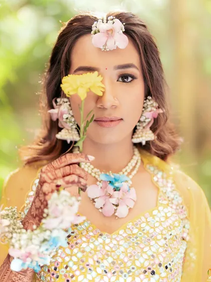 A creative portrait of the bride with a single yellow flower. Her custom floral maang tikka and necklace are visible, adding to the artistic feel.