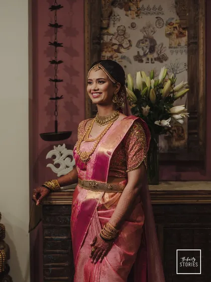 An elegant bridal portrait, with the bride standing gracefully in her pink silk saree. The traditional decor in the background adds to the classic feel of the image.