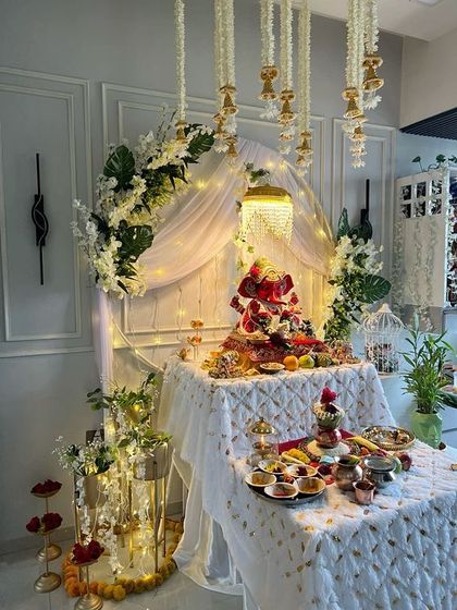 An elegant white and gold themed Ganpati setup. The decor features a white fabric canopy, hanging crystal strings, and a beautiful fruit arrangement on the idol, creating a look of pure sophistication.