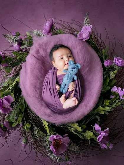 A beautifully styled newborn portrait. The baby is swaddled in purple and placed in a rustic nest adorned with matching flowers, holding a tiny blue teddy bear.