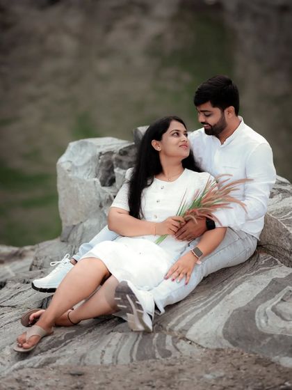 A couple sits on a rock, sharing a quiet and intimate moment, their white outfits contrasting with the natural stone.
