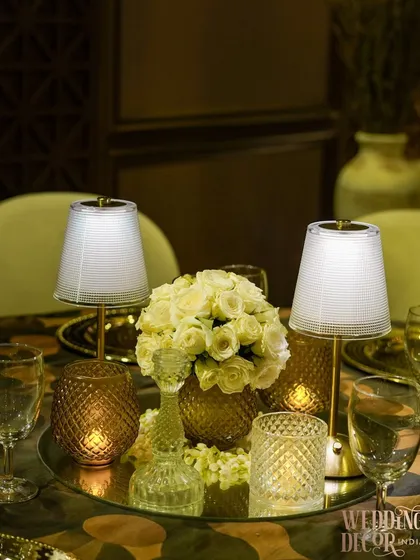 An elegant tabletop setup. A mirrored base reflects the light from the candles and miniature lamps, creating a sparkling effect around the central bouquet of white roses.
