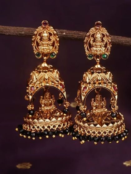 A close-up of the traditional temple jhumkis with a matte gold finish and black bead drops. The intricate detailing on the Lakshmi motif is clearly visible.